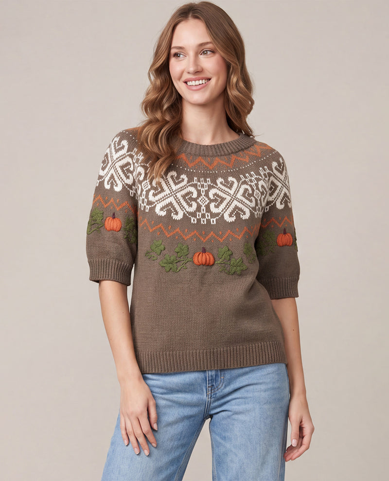 A woman wearing a brown knit sweater with white and orange Fair Isle style patterns and embroidered pumpkins, paired with blue jeans, against a solid beige background.