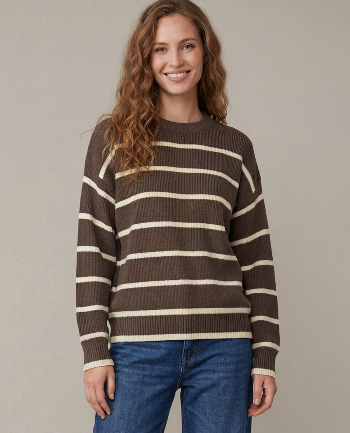 A woman wearing a brown and cream striped knit sweater with blue jeans, standing against a plain beige studio background.