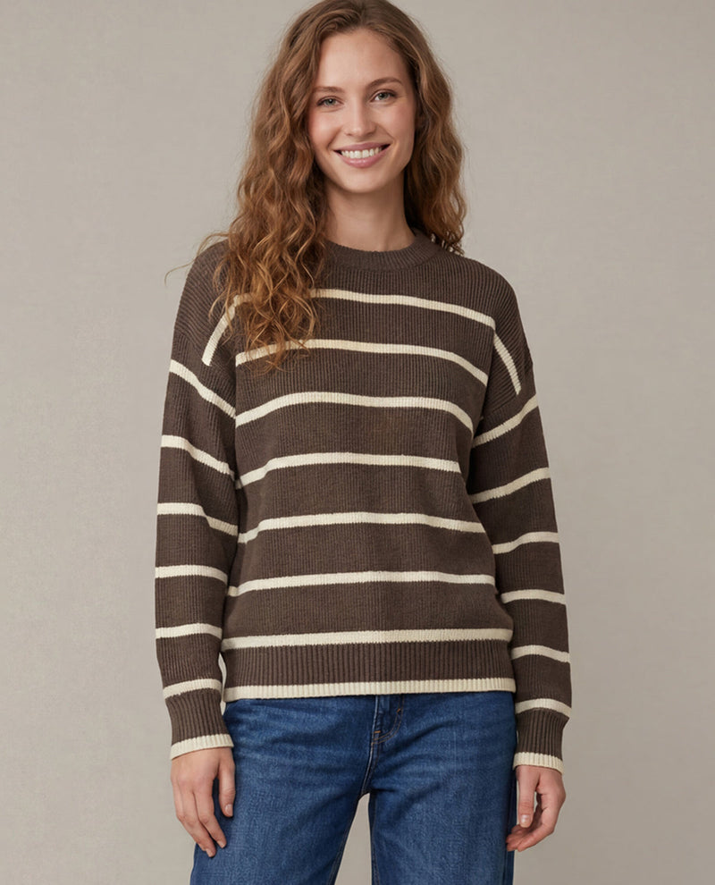 A woman wearing a brown and cream striped knit sweater with blue jeans, standing against a plain beige studio background.