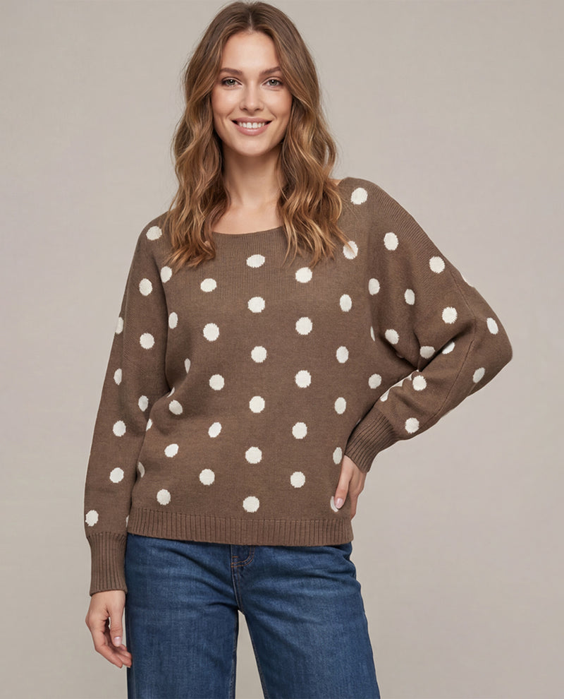 A woman wearing a brown knit sweater with large white polka dots and blue jeans, standing against a plain beige studio background.