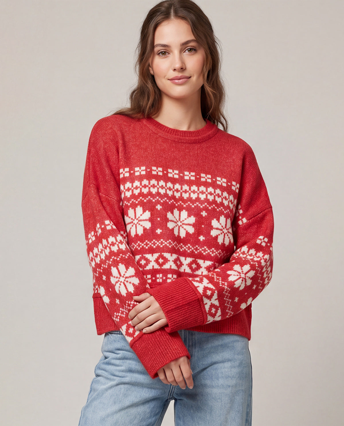 A woman wearing a red crewneck sweater with a white Nordic/Fair Isle snowflake pattern, paired with light blue jeans, against a neutral beige background.