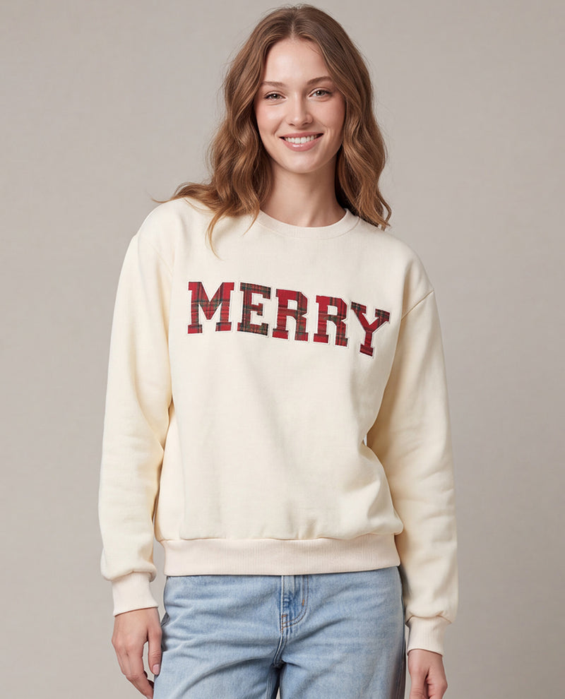 A woman wearing a cream-colored crewneck sweatshirt with 'MERRY' printed across the chest in red and black plaid letters, paired with light blue jeans, against a neutral beige background.
