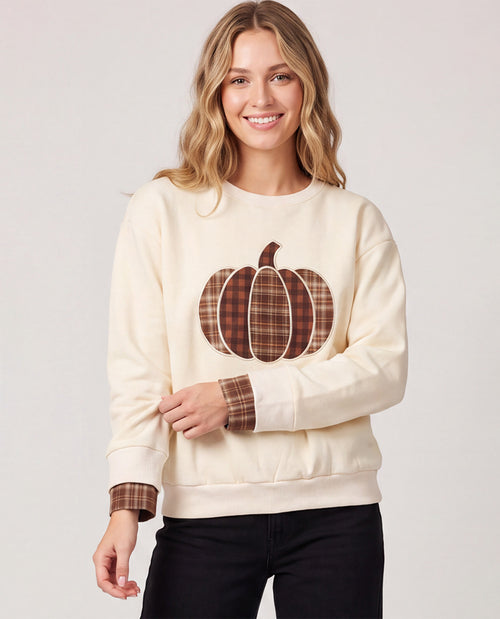 A woman wearing a cream-colored crewneck sweatshirt with a brown and red plaid pumpkin graphic on the front and matching plaid cuff details, paired with black pants, against a light gray studio background.