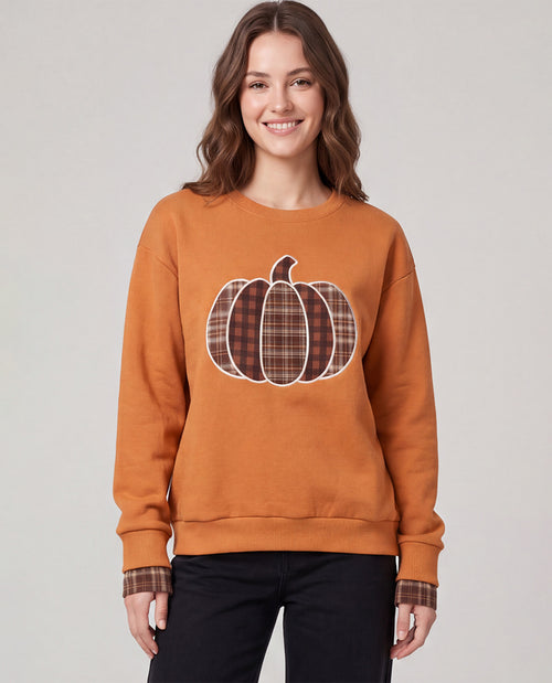 A woman wearing a rust-orange crewneck sweatshirt with a plaid pumpkin graphic on the front, paired with black pants, against a solid light gray background.