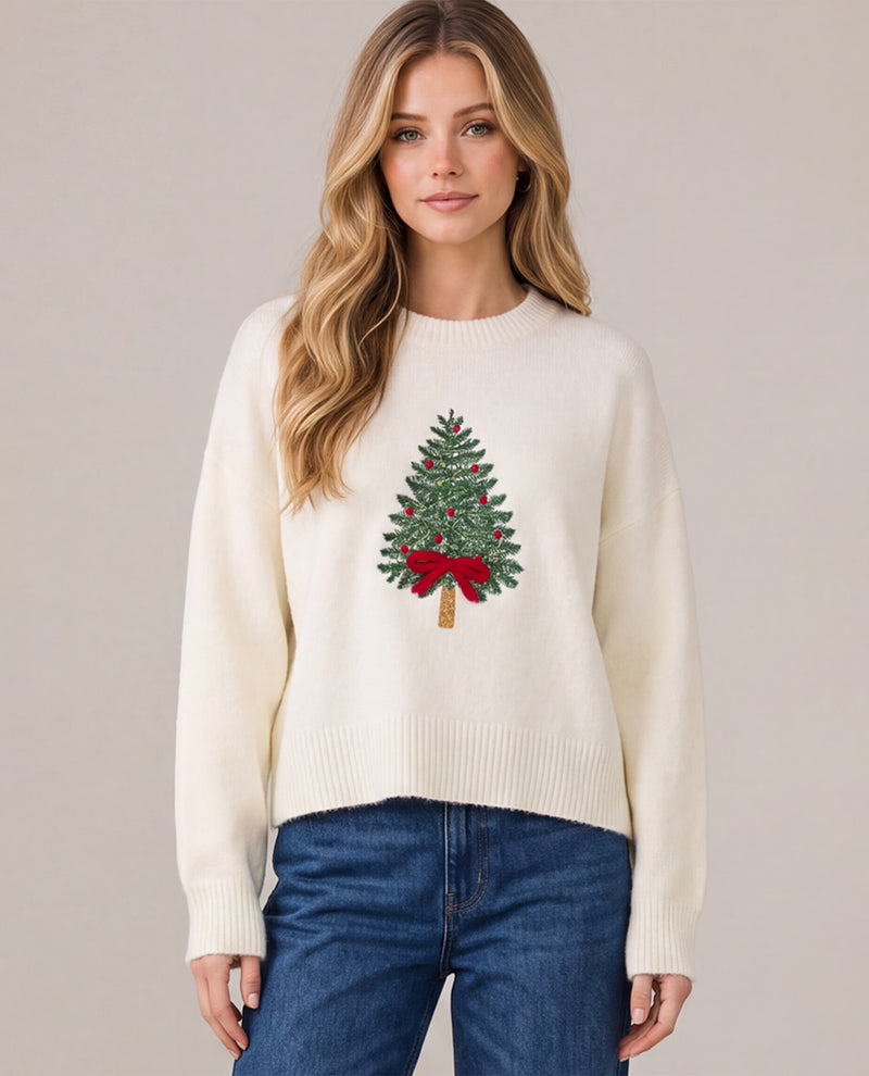 A woman wearing a cream-colored knit sweater with an embroidered green Christmas tree and red bow on the front, paired with blue jeans, against a solid light gray background.
