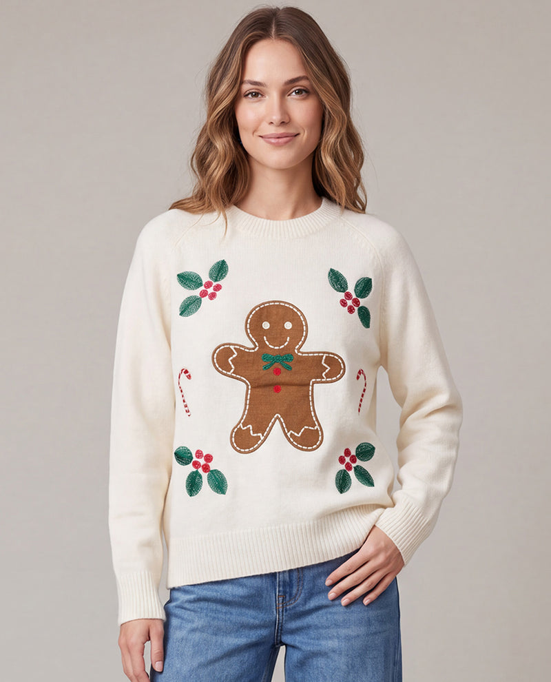 A woman wearing a cream-colored crewneck sweater with a brown gingerbread man design, green holly leaves, and red berries, paired with blue jeans, against a solid light gray background.