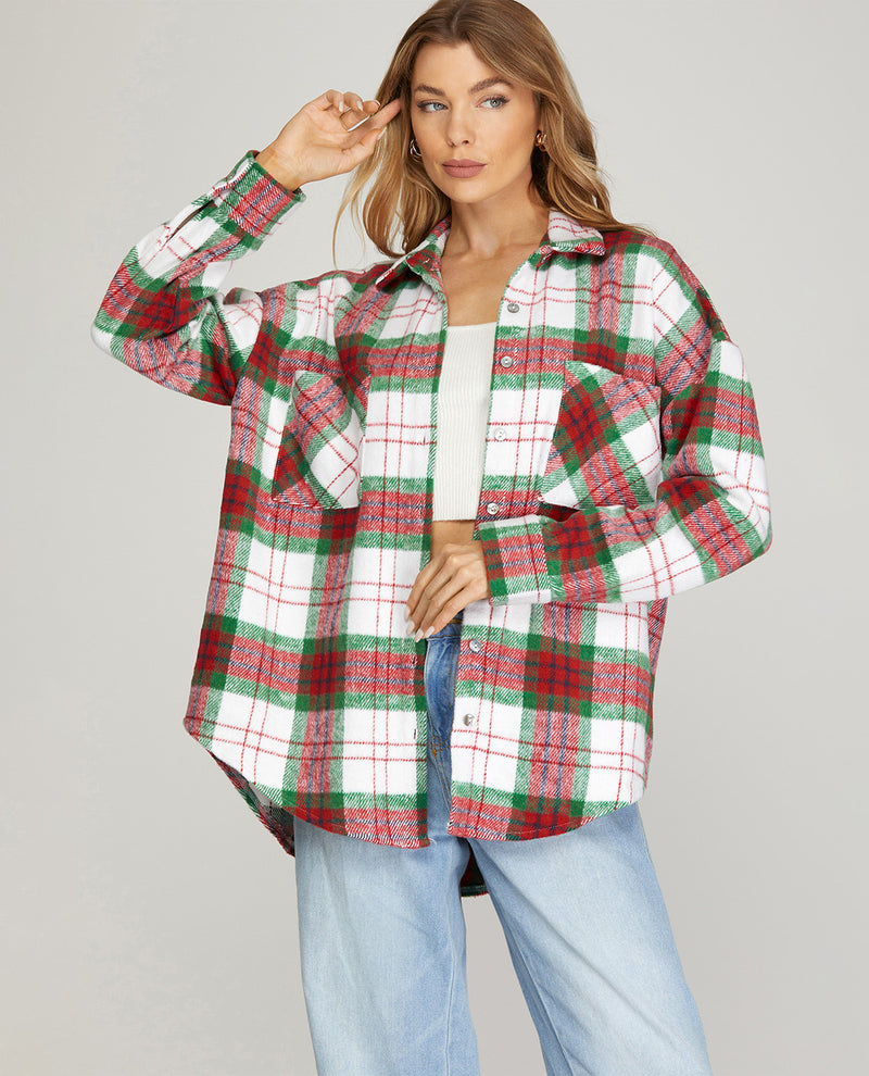 A woman modeling a long-sleeve button-up plaid shirt in red, green, and white, worn open over a white crop top and light blue jeans, against a solid light gray background.