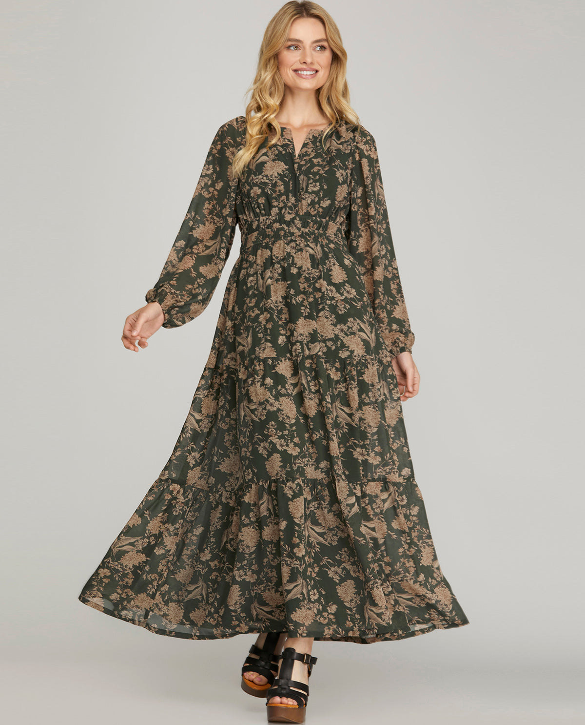 A woman modeling a long-sleeved, floor-length maxi dress with a dark green and beige floral pattern, paired with black platform sandals, against a solid light gray background.