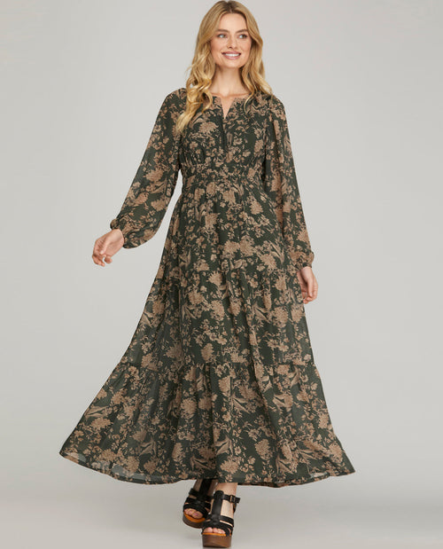 A woman modeling a long-sleeved, floor-length maxi dress with a dark green and beige floral pattern, paired with black platform sandals, against a solid light gray background.