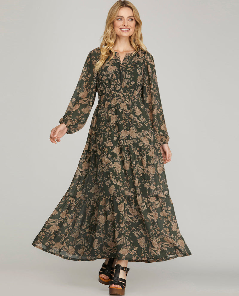 A woman modeling a long-sleeved, floor-length maxi dress with a dark green and beige floral pattern, paired with black platform sandals, against a solid light gray background.