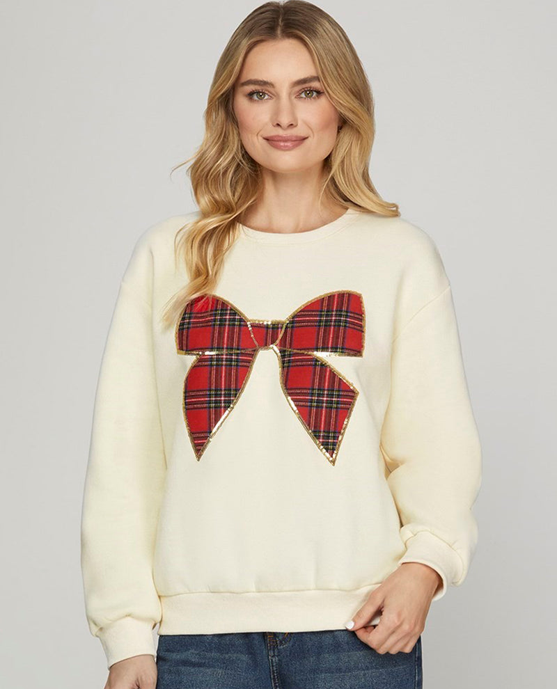 Model wearing a cream color long sleeve top with a red plaid bow on the front 