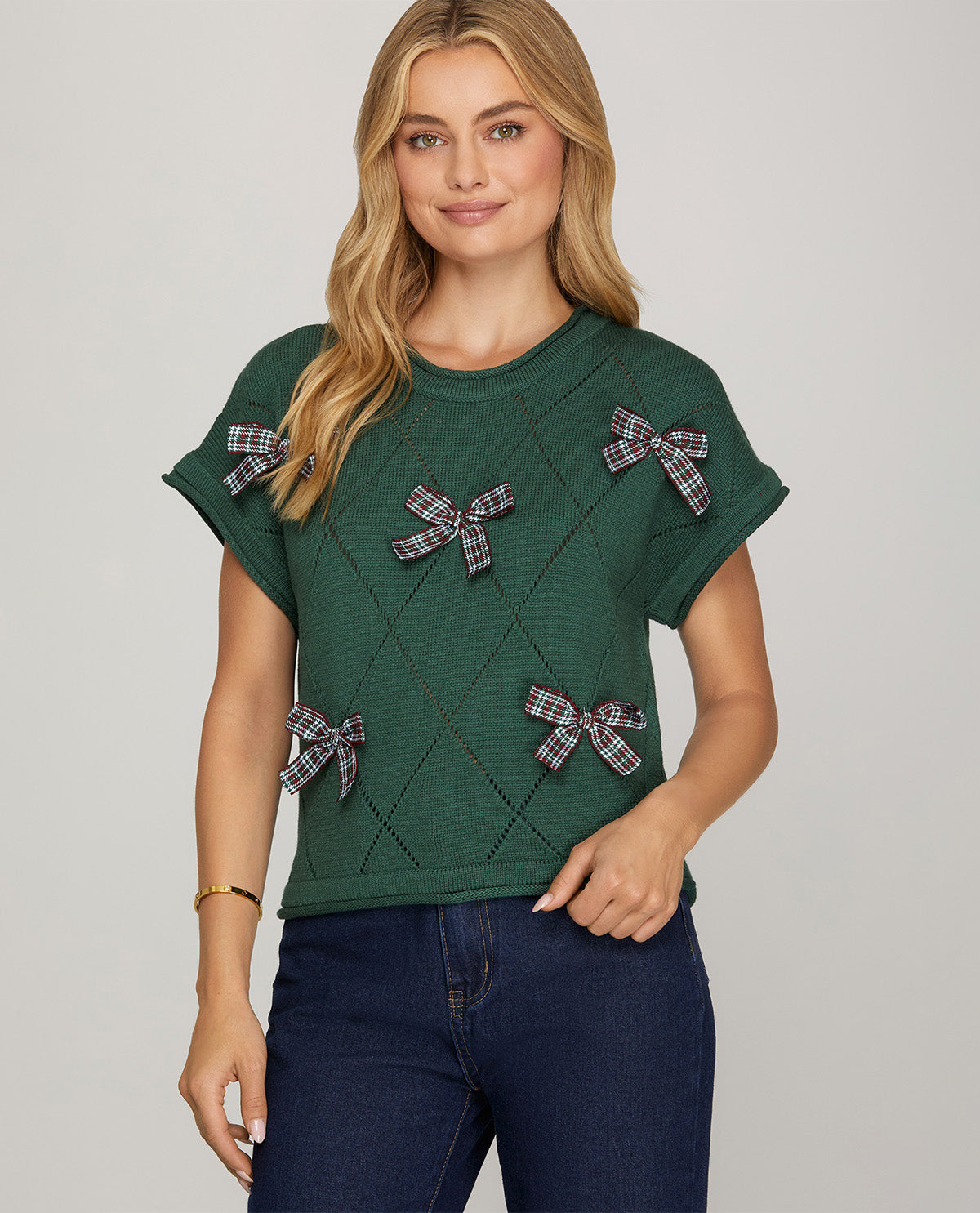A woman modeling a dark green short-sleeve knit top with a diamond argyle pattern and plaid bow accents, paired with dark blue jeans against a light gray studio background.