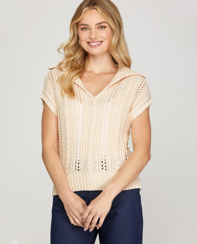 A woman wearing a cream-colored knit short-sleeve top with a hood and dark blue jeans, standing against a light gray studio background.