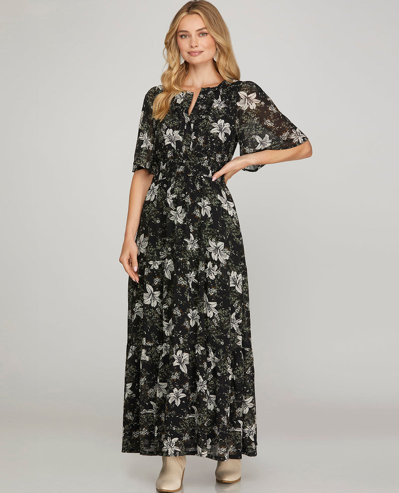 A woman modeling a black midi dress with a white floral pattern, short flutter sleeves, and a split neckline, photographed against a solid light gray background.