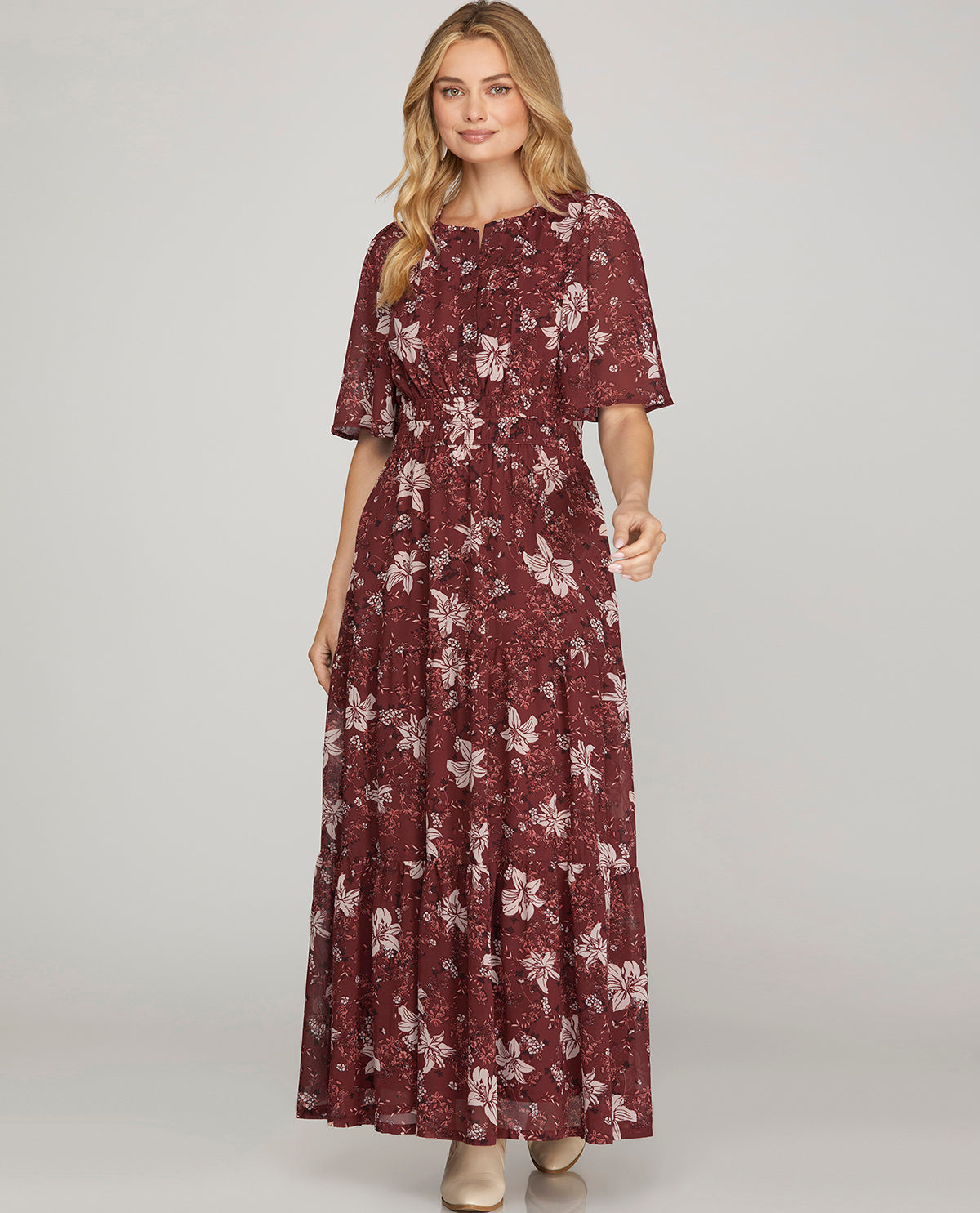 A woman modeling a floor-length, short-sleeve dress with a dark red floral pattern against a light gray studio background.