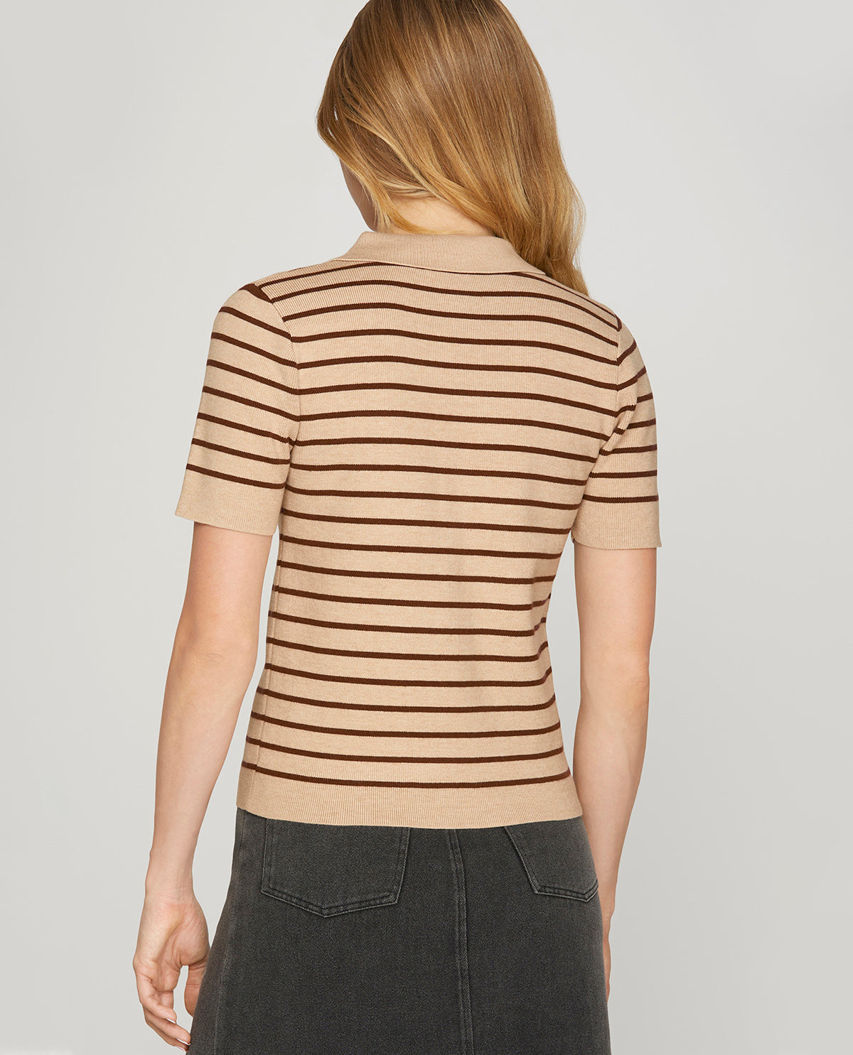 A woman shown from the back wearing a beige short-sleeve knit top with thin dark brown horizontal stripes, paired with a dark gray skirt, against a light gray studio background.