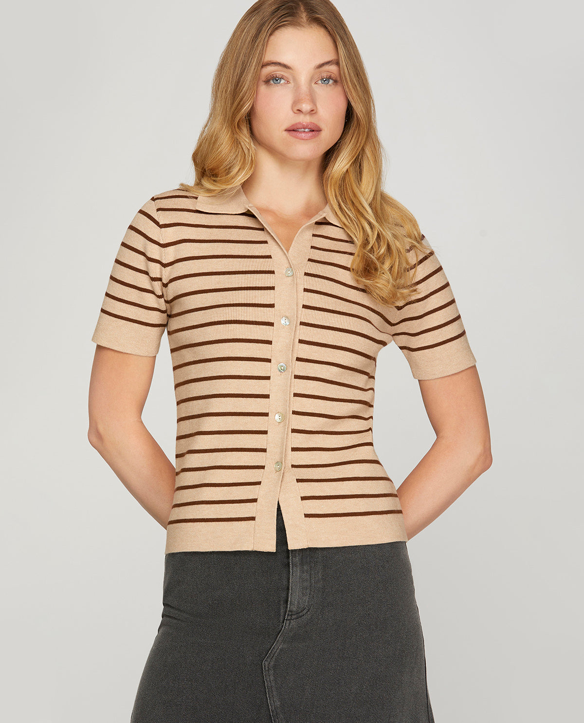 A woman modeling a beige short-sleeve knit polo shirt with thin dark brown horizontal stripes, paired with dark gray jeans, against a solid light gray background.