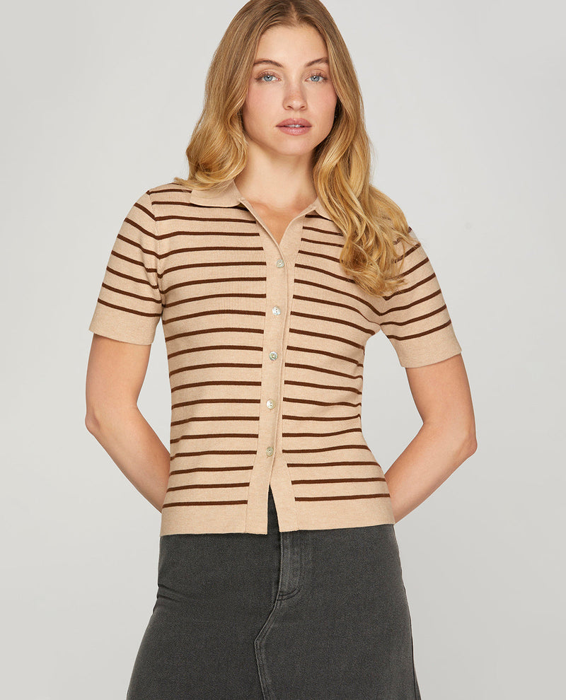 A woman modeling a beige short-sleeve knit polo shirt with thin dark brown horizontal stripes, paired with dark gray jeans, against a solid light gray background.