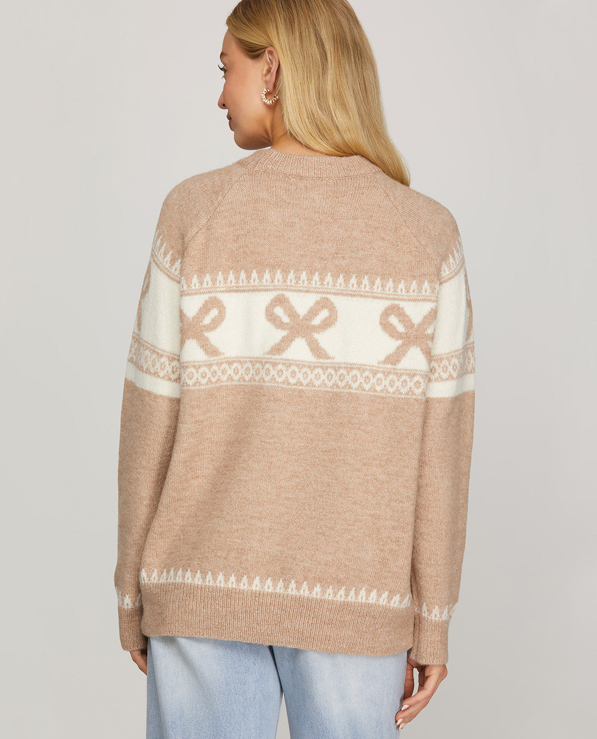 A woman shown from the back wearing a beige knit sweater with a cream-colored horizontal pattern band featuring bow motifs and geometric borders, paired with light blue jeans against a solid light gray background.