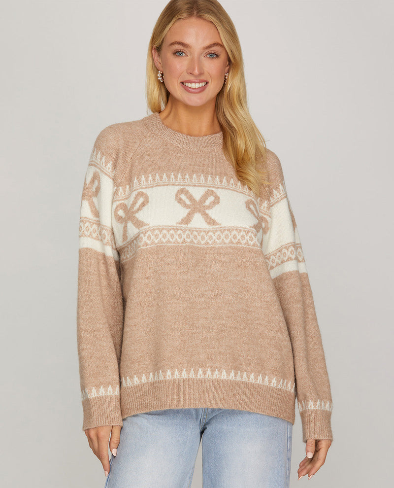 A woman wearing a beige crewneck knit sweater with a cream Nordic bow pattern, paired with light blue jeans, against a solid light gray background.