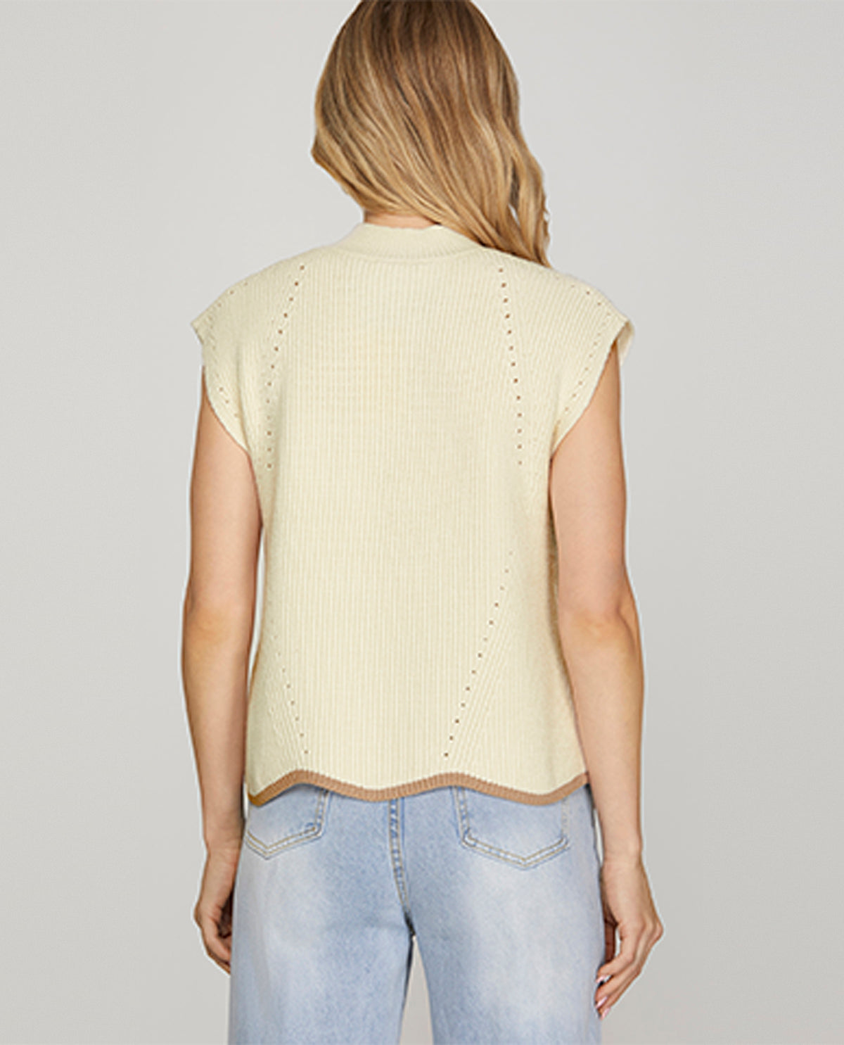 Woman wearing a light yellow sleeveless sweater and light blue jeans on a plain background