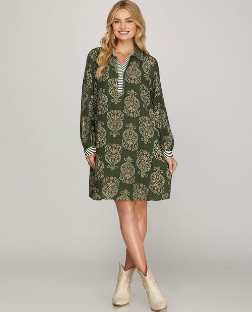 A woman modeling a dark green long-sleeve collared dress with a beige and white ornamental pattern, styled with a striped shirt underneath and beige ankle boots, against a solid light gray background.