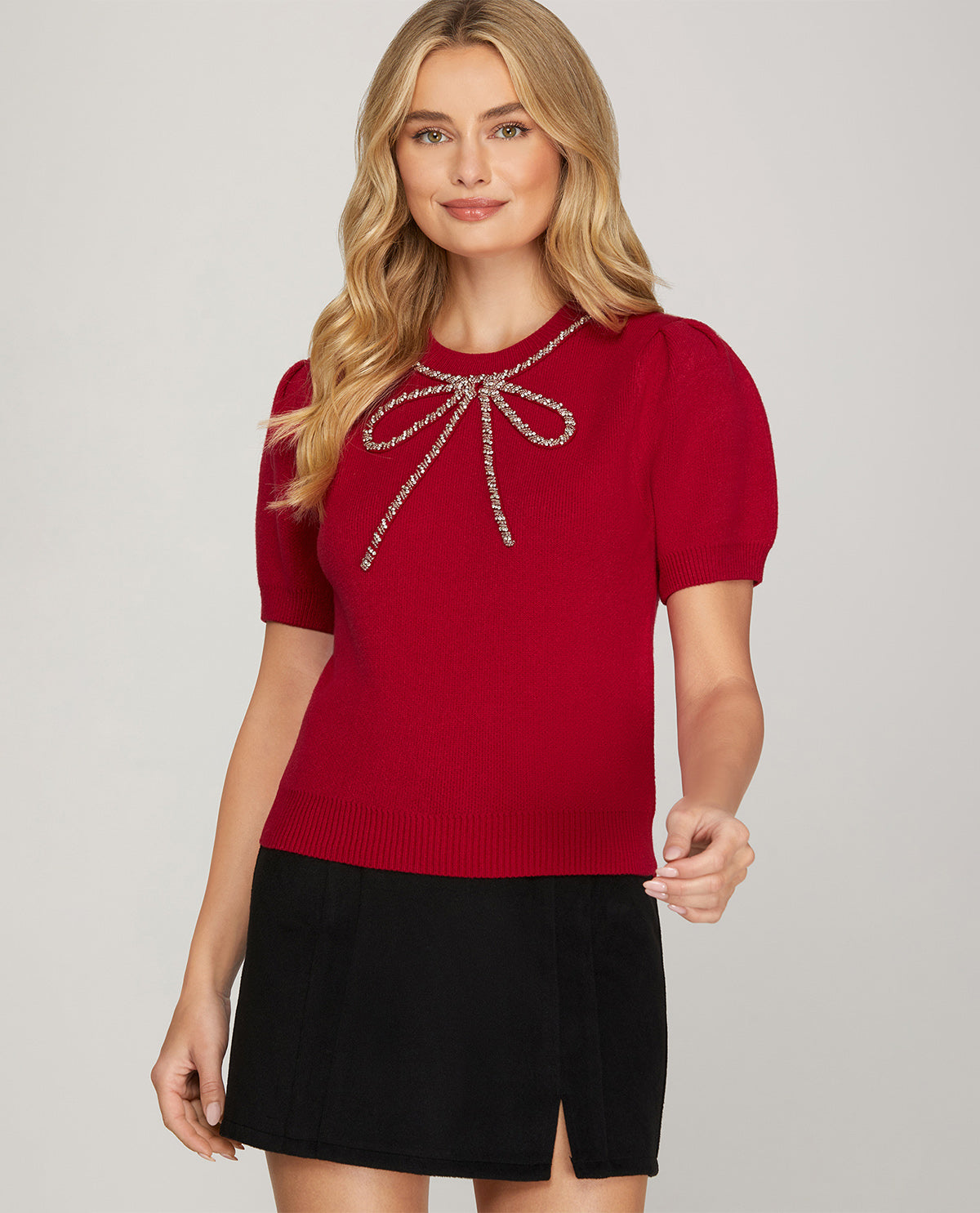 A woman modeling a short-sleeve red knit top with a rhinestone bow detail at the neckline, paired with a black skirt, against a light gray studio background.