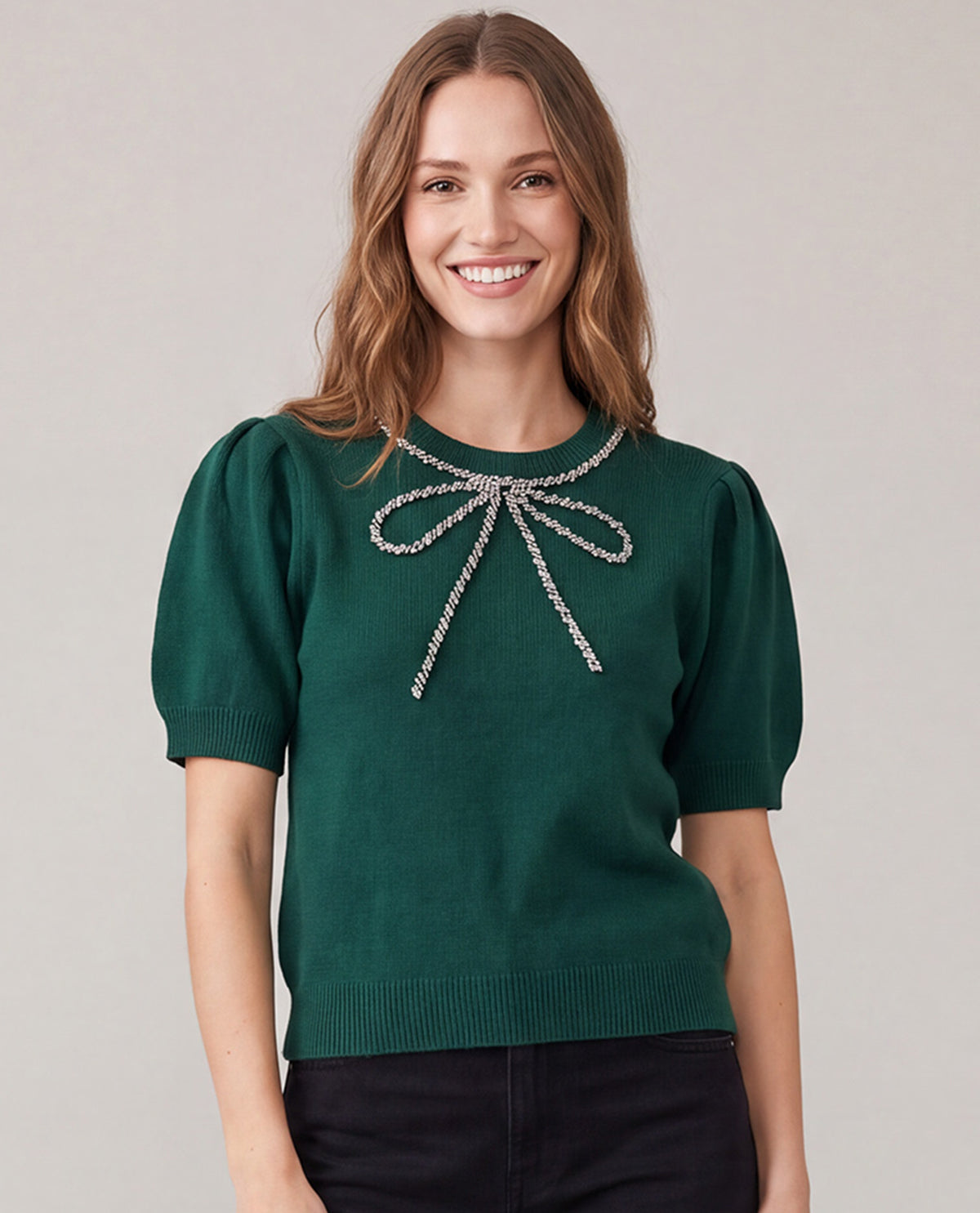 A woman wearing a dark green short-sleeve knit sweater with a silver rhinestone bow detail at the neckline, paired with black pants, against a solid light gray background.