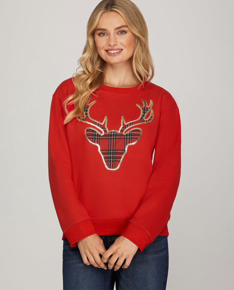 A woman wearing a red crewneck sweatshirt with a large plaid reindeer graphic on the front, paired with dark blue jeans, against a plain light gray studio background.