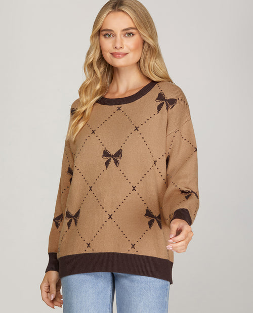 A woman modeling a brown knit sweater with a darker brown diamond lattice pattern and bow motifs, paired with light blue jeans against a light gray studio background.