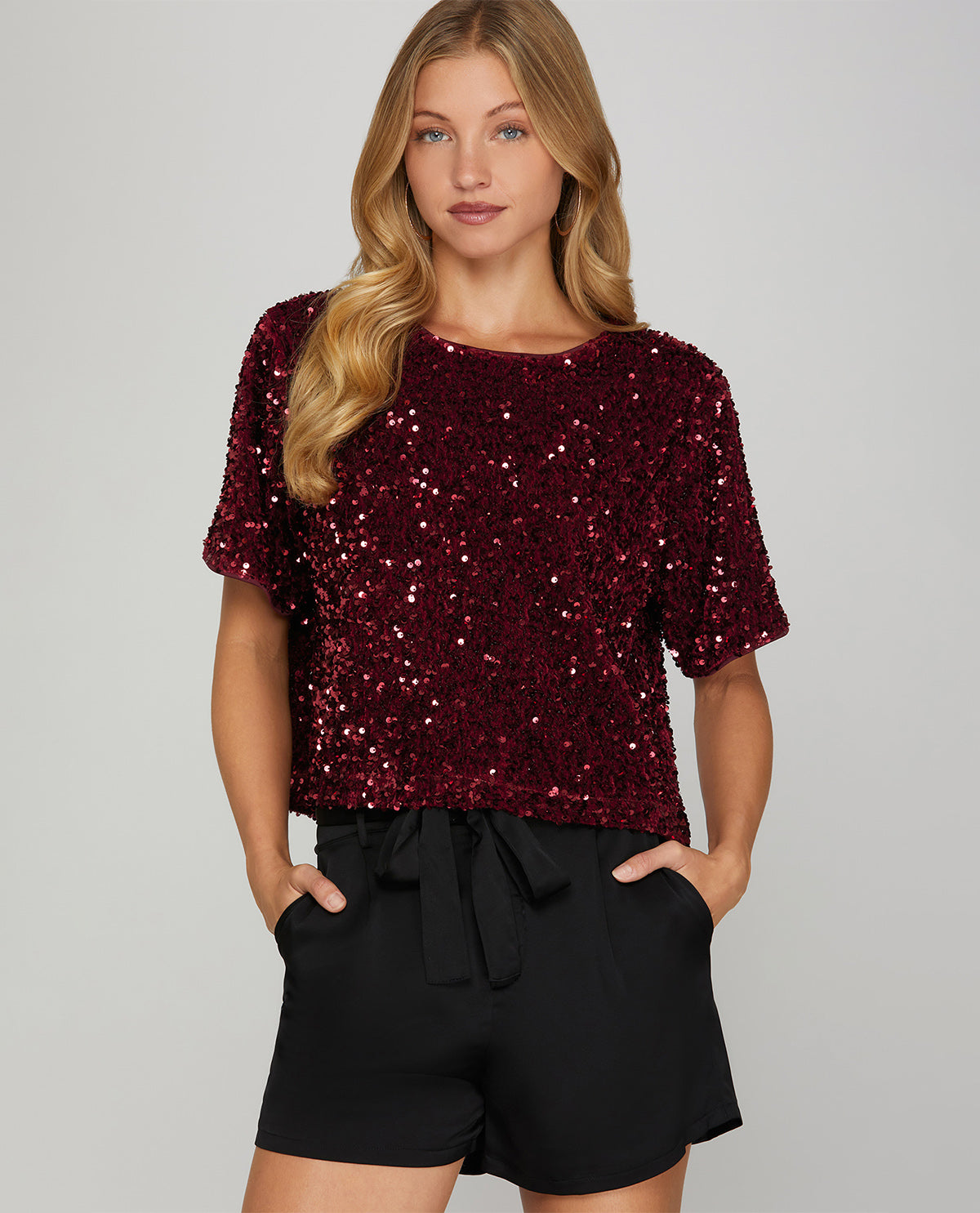 A woman modeling a short-sleeve burgundy sequined top paired with black shorts against a light gray studio background.