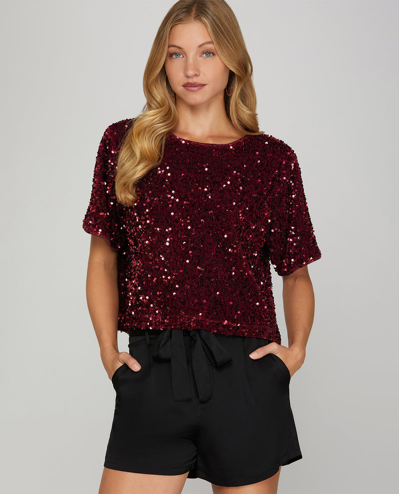 A woman modeling a short-sleeve burgundy sequined top paired with black shorts against a light gray studio background.