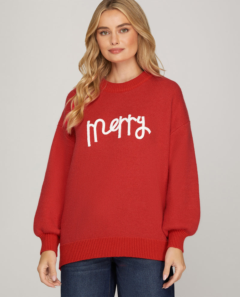 A woman wearing a red crewneck sweater with 'Merry' written in white script across the chest, paired with dark blue jeans, against a solid light gray background.