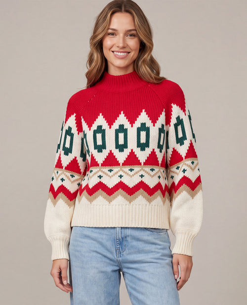 A woman wearing a red, cream, and green Fair Isle knit sweater with light blue jeans, standing against a solid beige studio background.