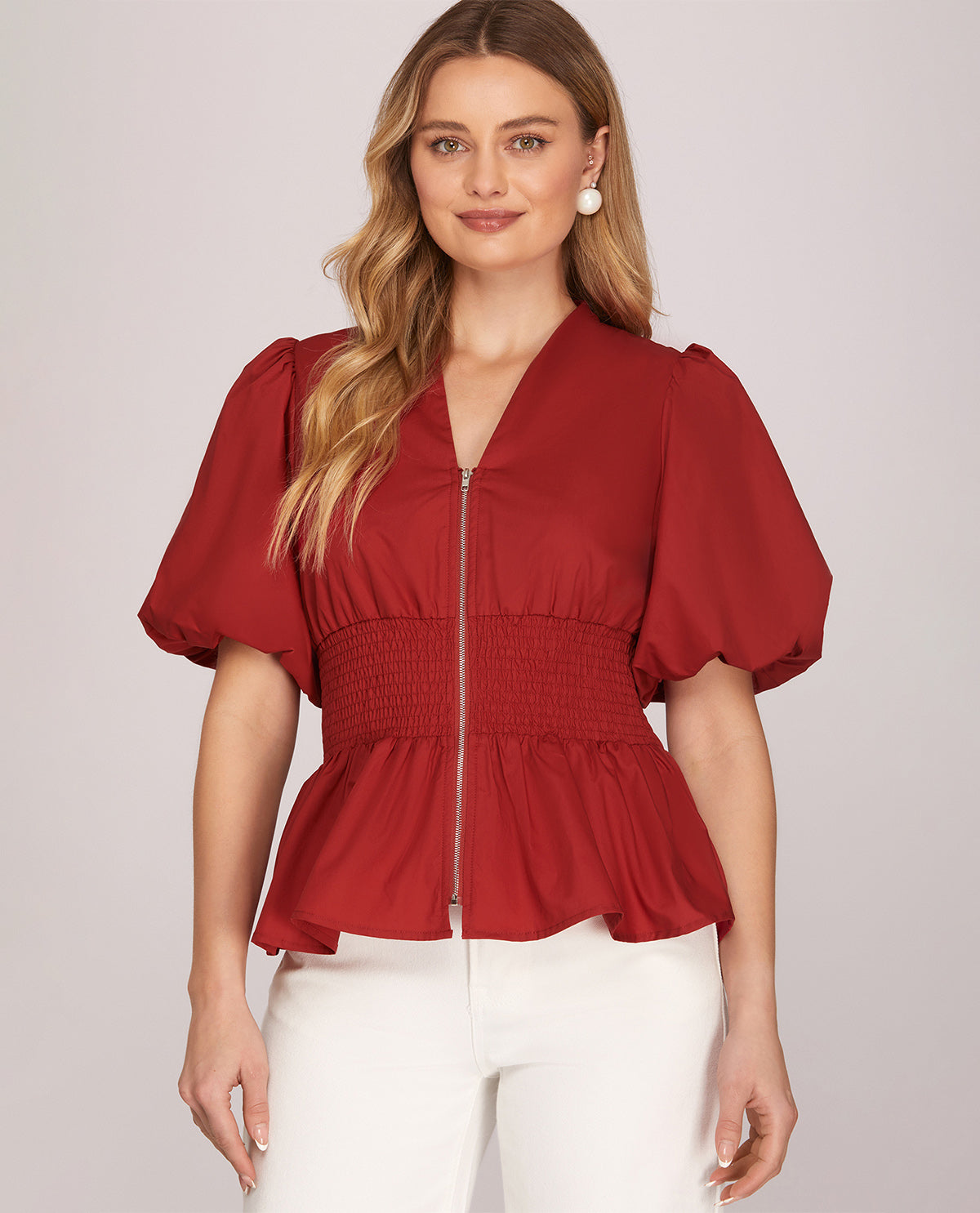 A woman wearing a red short-sleeve blouse with puff sleeves, a smocked waist, and a front zipper, paired with white pants, against a light gray studio background.