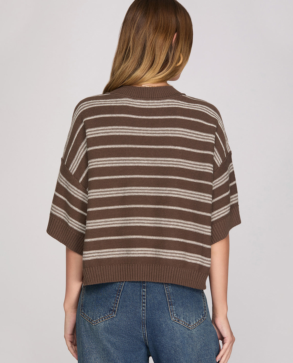A woman viewed from the back wearing a brown and beige horizontally striped knit sweater with elbow-length sleeves, paired with blue jeans, against a light gray studio background.