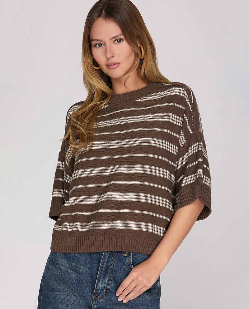 A woman wearing a brown and beige horizontally striped knit sweater with elbow-length sleeves, paired with blue jeans, against a light gray studio background.