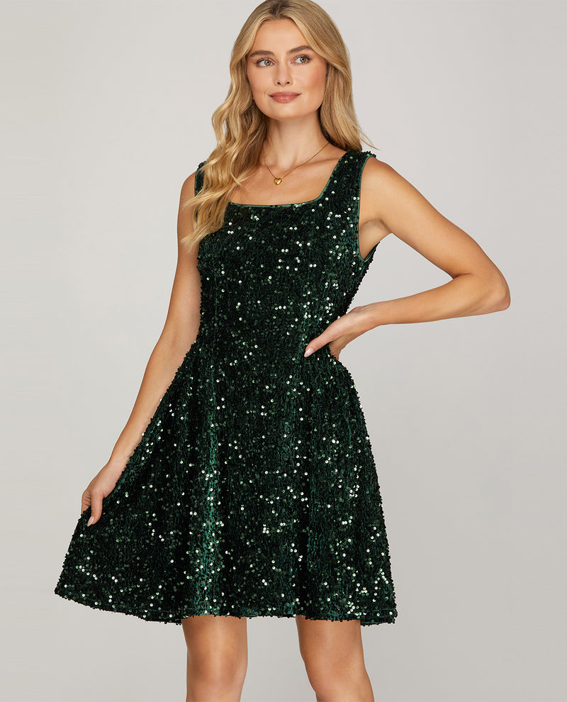 A woman modeling a sleeveless, knee-length dark green sequined dress against a plain light gray studio background.