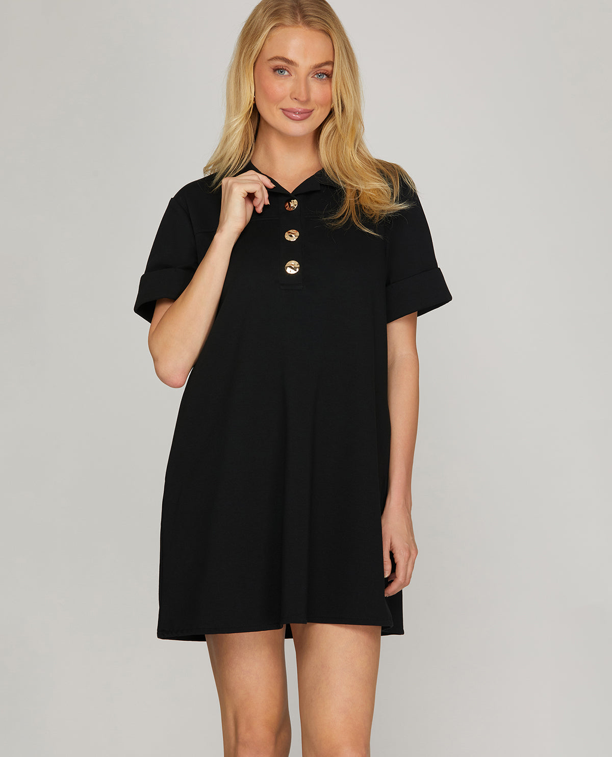 A blonde woman modeling a short-sleeve black polo dress with gold buttons on the chest, photographed against a neutral light gray studio background.