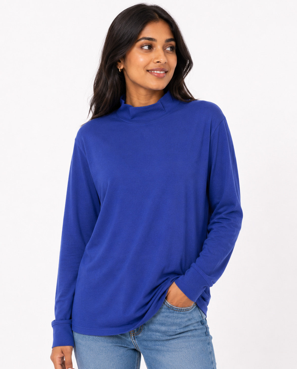 A woman wearing a royal blue long-sleeve mock neck top and light wash blue jeans, standing against a plain white studio background.