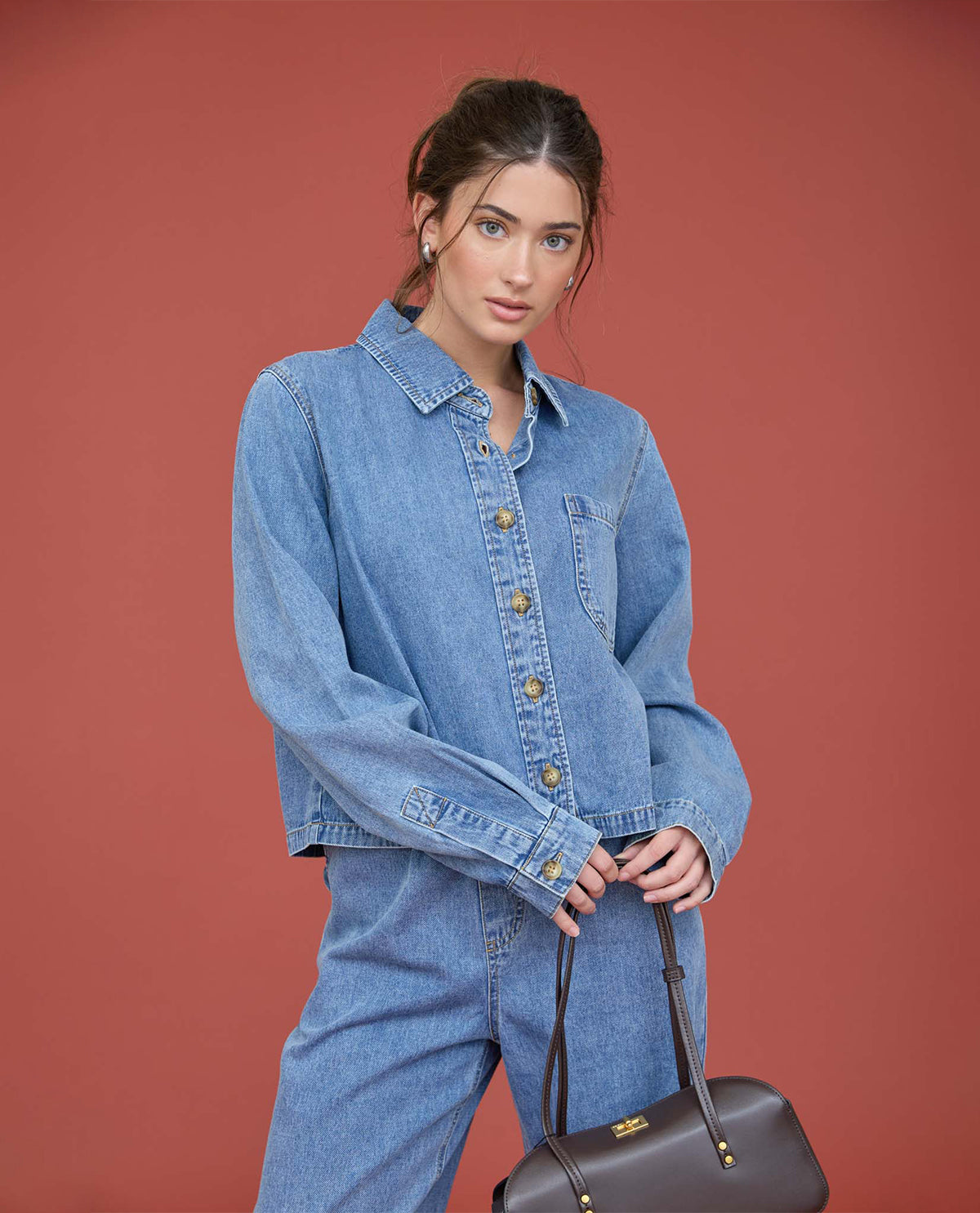 A woman modeling a light blue denim jacket and matching denim pants against a solid terracotta background, holding a dark brown leather handbag.