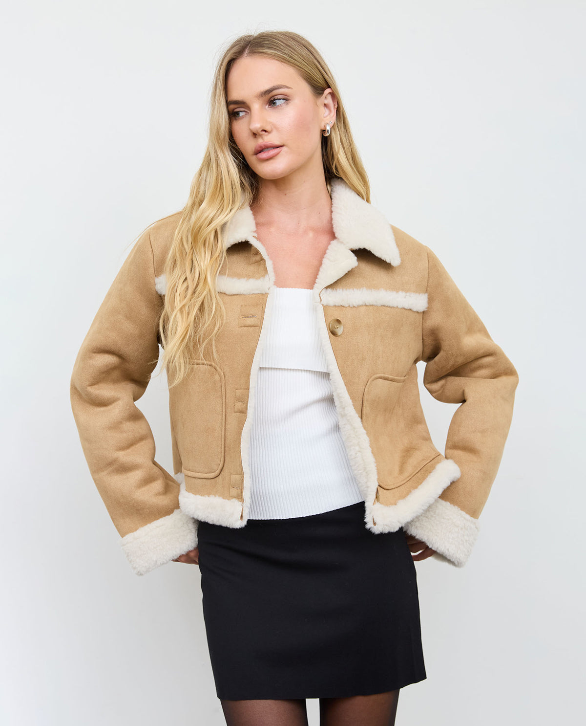 A blonde model wearing a cropped tan shearling jacket with a white ribbed collar, cuffs, and hem, styled over a white top and a black skirt, against a light gray studio background.