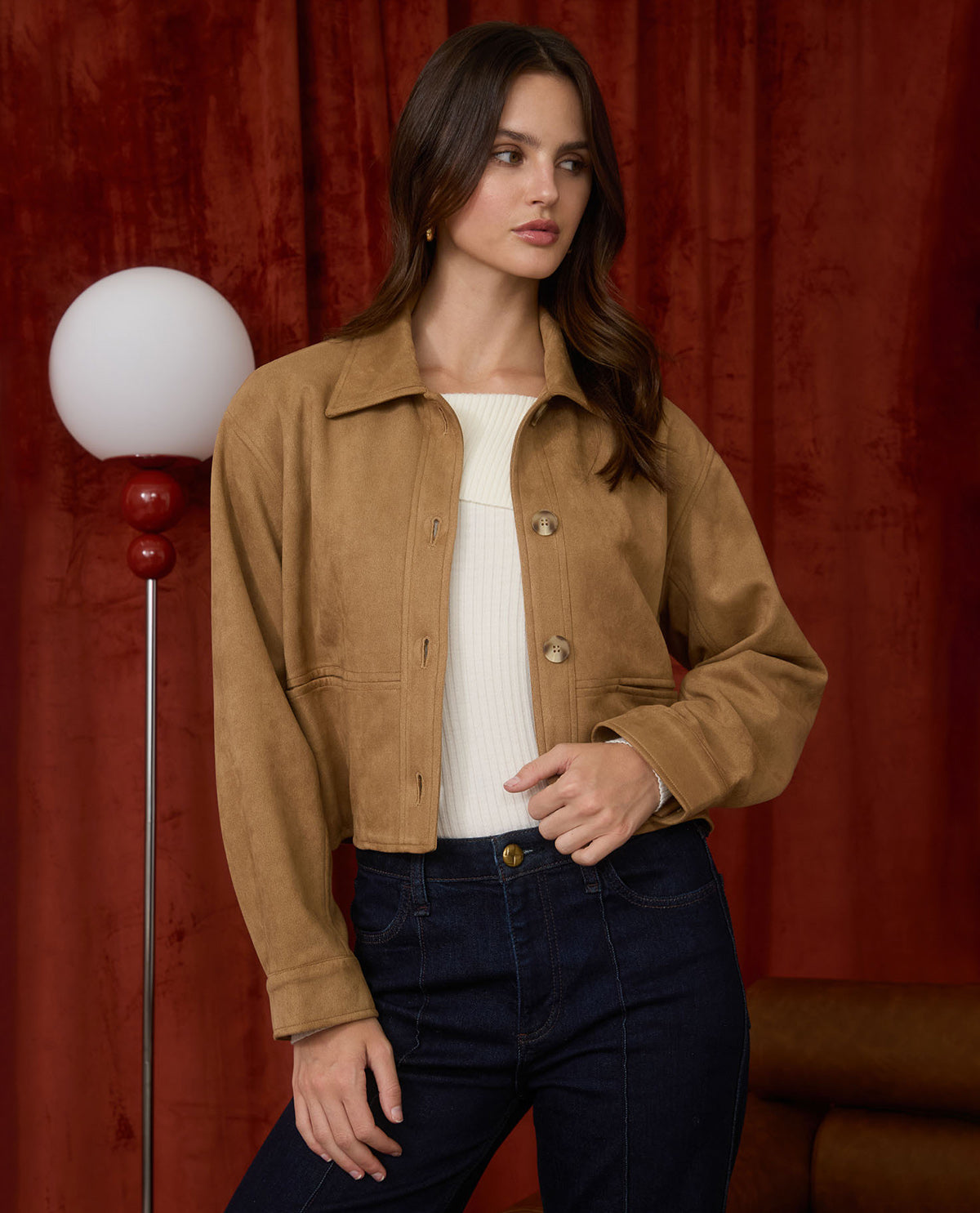 A woman models a cropped tan suede jacket over a white ribbed top and dark blue jeans, standing in front of a dark red velvet curtain with a vintage-style floor lamp on the left.