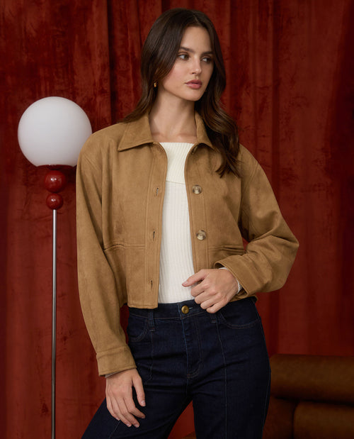 A woman models a cropped tan suede jacket over a white ribbed top and dark blue jeans, standing in front of a dark red velvet curtain with a vintage-style floor lamp on the left.