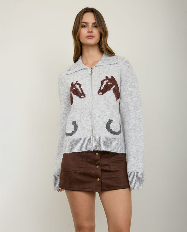 A woman models a light gray knit zip-up cardigan featuring brown horse head motifs and dark gray horseshoes, paired with a brown mini skirt against a plain light gray studio background.