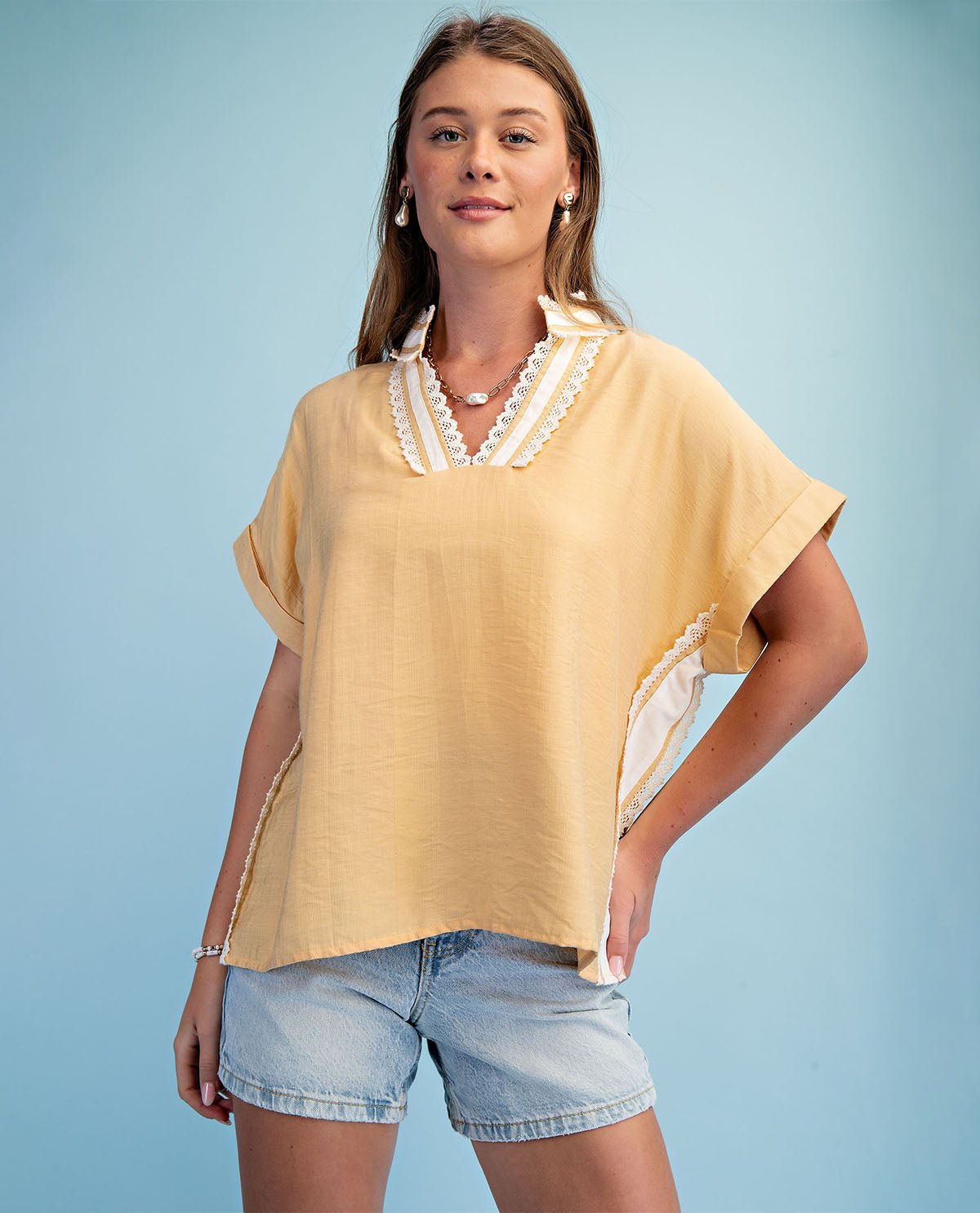 Woman wearing a yellow top with lace details and denim shorts against a light blue background
