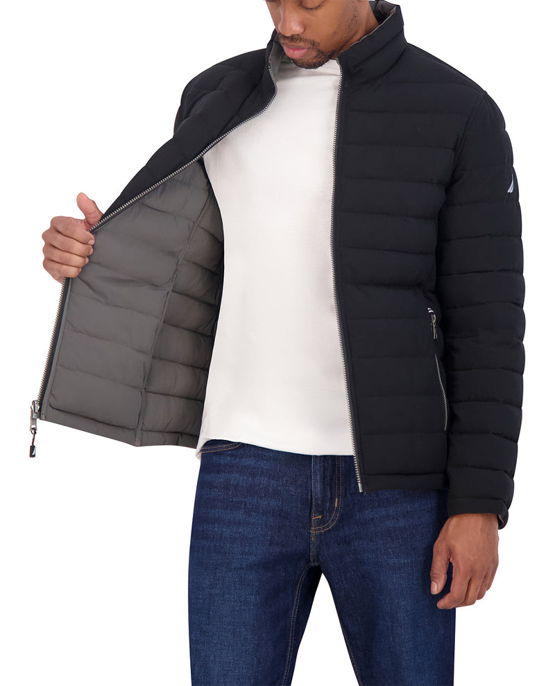 A man wearing a black puffer jacket with a gray interior lining, shown from the chest down against a white background. He is wearing a white t-shirt and blue jeans, and is holding the jacket open to show the lining.