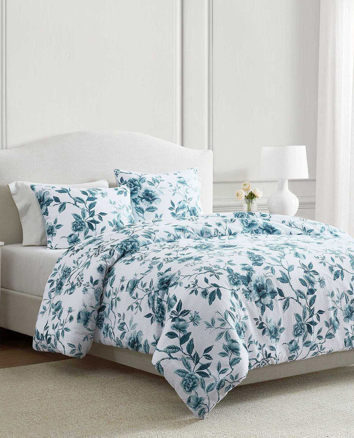 A bed dressed in a white bedding set with a teal floral and leaf pattern, styled in a bright bedroom with a white upholstered headboard, bedside lamp, and small vase of flowers.