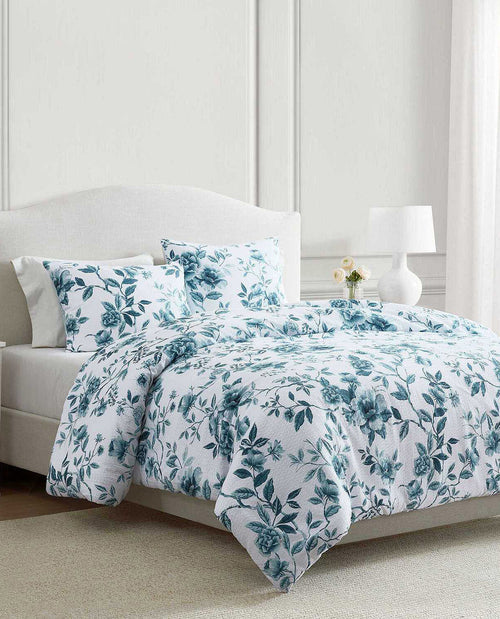 A bed dressed in a white bedding set with a teal floral and leaf pattern, styled in a bright bedroom with a white upholstered headboard, bedside lamp, and small vase of flowers.