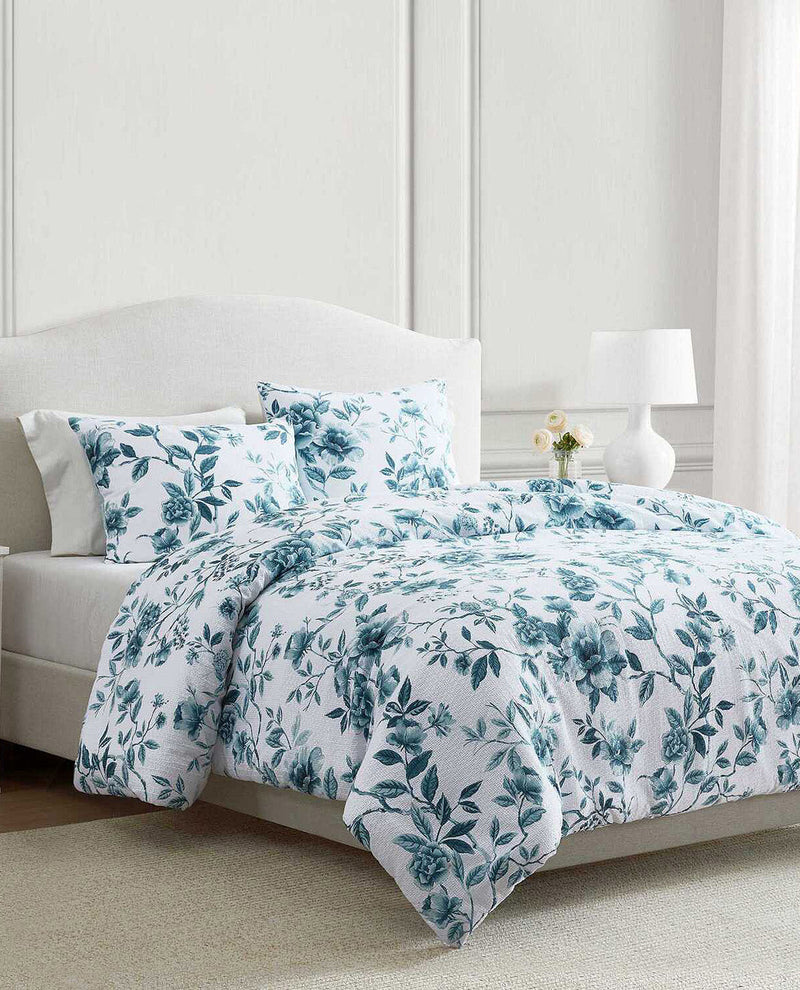 A bed dressed in a white bedding set with a teal floral and leaf pattern, styled in a bright bedroom with a white upholstered headboard, bedside lamp, and small vase of flowers.